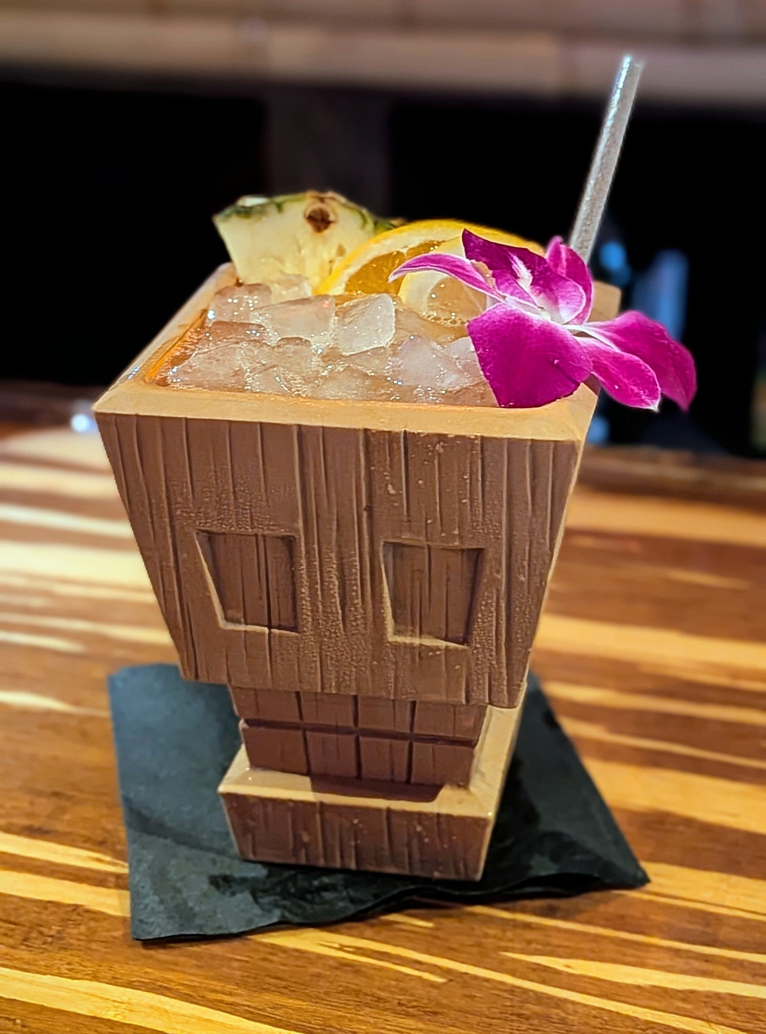 Picture of a ceramic skull tiki mug with wood grain full of ice with fruit and a straw ready to drink.