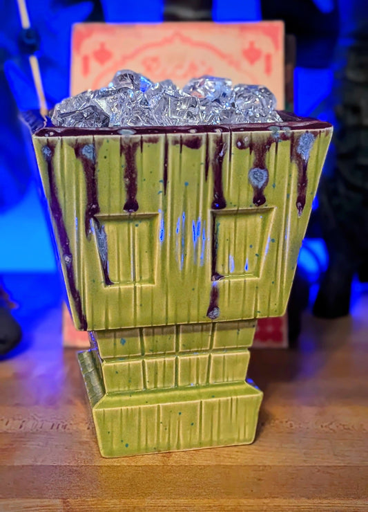 Green Apple speckled skull tiki mug with purple glaze drips on the top edges with a wood grain texture. 