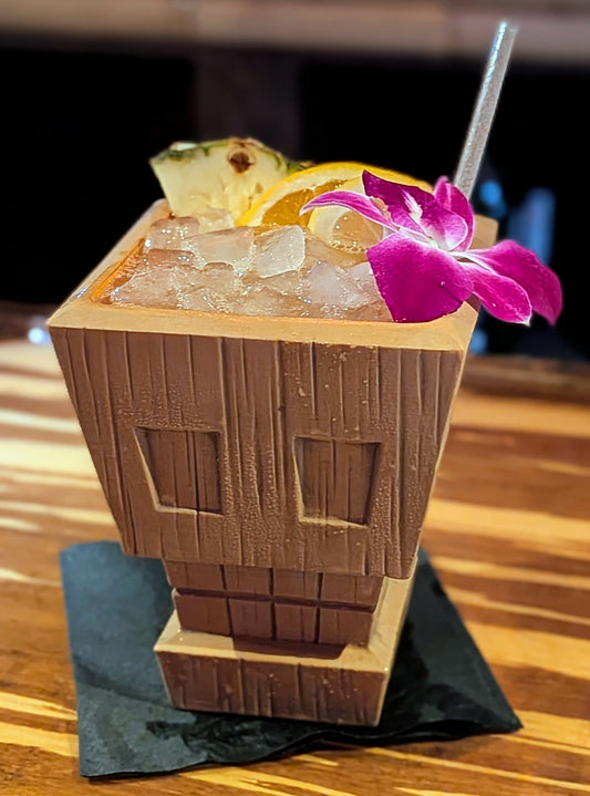 Tiki-style cocktail with pineapple and flower garnish in a wooden cup on a wooden surface.